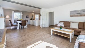 Amplia sala de estar con piso de madera, un sofá marrón, una mesa de centro blanca y un comedor con sillas y una mesa blancos.