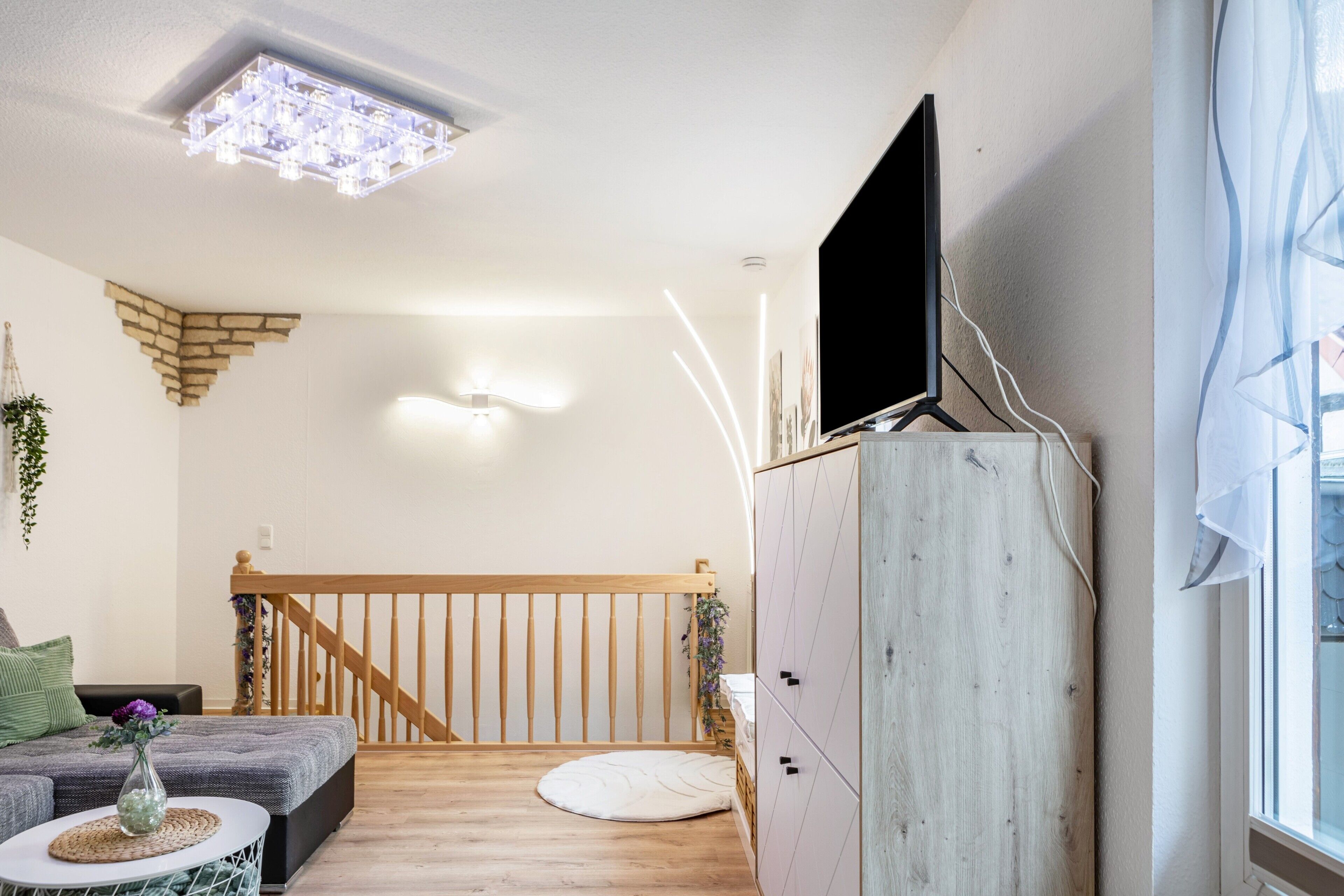 Una habitación con una escalera de madera, un televisor de pantalla plana montado sobre un mueble de madera, un tapete blanco redondo y una mesita redonda con un jarrón.