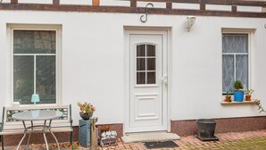 Un edificio blanco con puerta principal, dos ventanas, un banco y plantas en macetas.