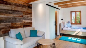 A room with a wooden wall, a sofa with teal cushions, a round wooden table, and a bed with patterned bedding. - Room 'Apartment Mit Bergblick' with Mountain View, Balcony and Wi-Fi (Krün)