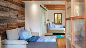 A cozy room with a wooden wall, a sofa with patterned cushions, a bed with a wooden headboard, and a window with a view of greenery. - Room 'Apartment Mit Bergblick' with Mountain View, Balcony and Wi-Fi (Krün)
