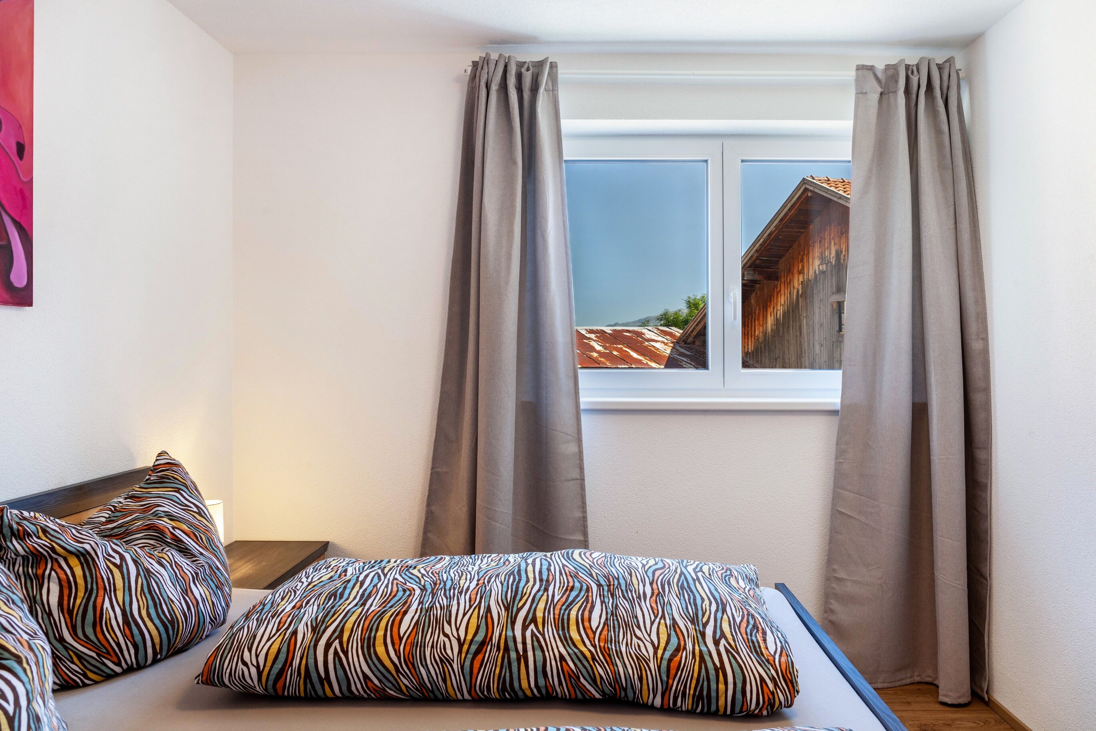 A bedroom with two beds, grey curtains, a window with a view of a building, and a painting on the wall.