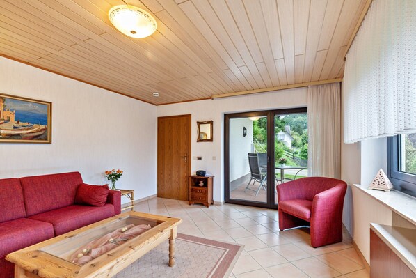 Un salon spacieux avec un canapé rouge, une table basse en bois et un fauteuil rouge. Une porte-fenêtre coulissante donne accès à un balcon aménagé avec des sièges d’extérieur.