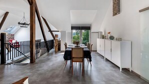 Une salle à manger spacieuse avec un plafond haut, des poutres apparentes et une vue sur l’extérieur grâce à de grandes fenêtres.