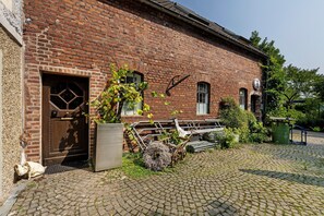 A brick building with a wooden door, a wheelbarrow, and a green trash bin.