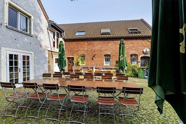 A courtyard with a large wooden table and chairs, green umbrellas, and a brick building with windows and a door.