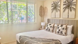 A bedroom with a large bed, a window with a view of greenery, and two wall-mounted palm tree paintings.