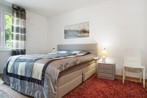 Ein Schlafzimmer mit einem Bett, einem Nachttisch, einem Stuhl, einer Lampe, einem Bild und einem Fenster mit Vorhängen.