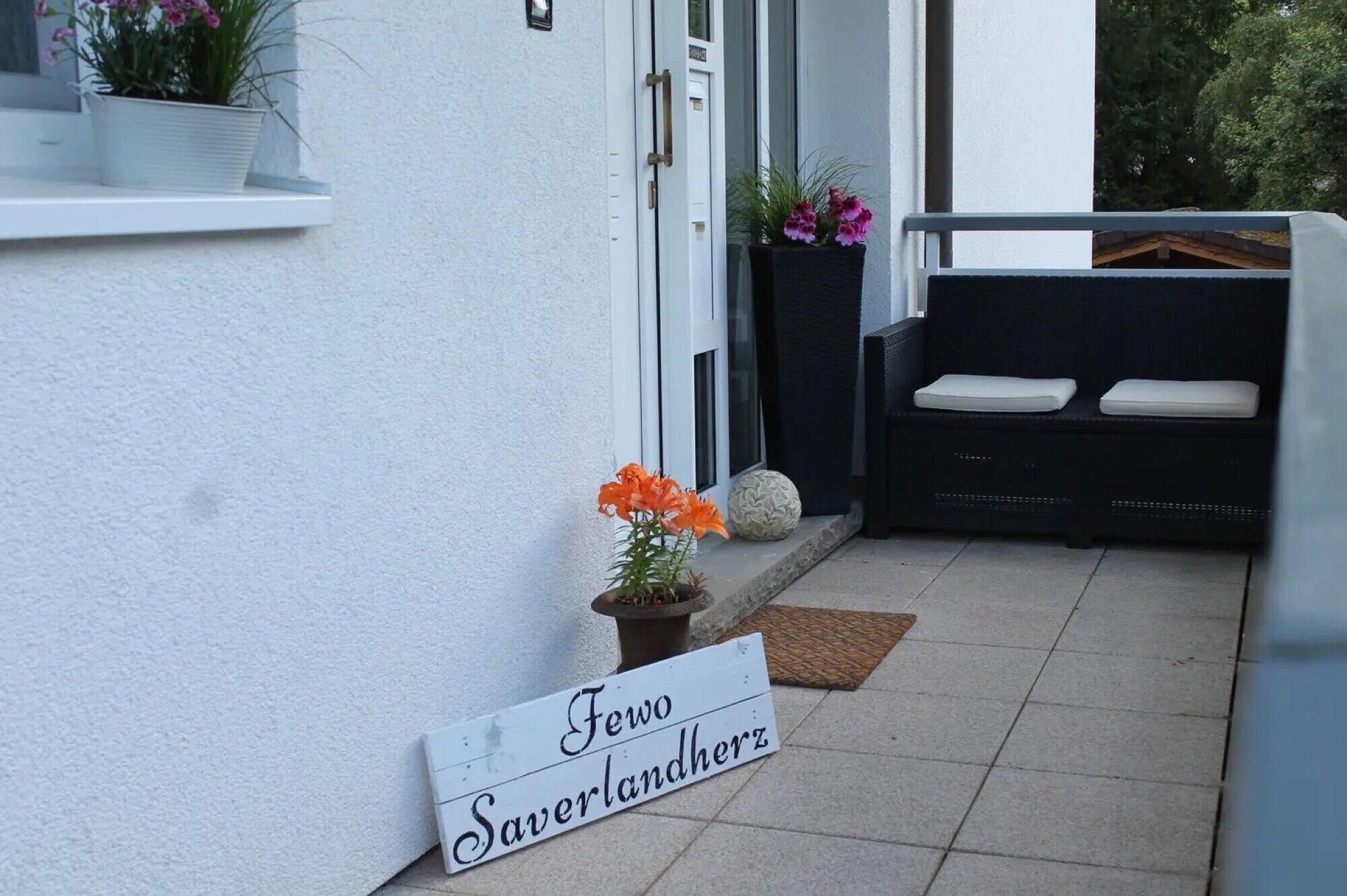 Eine weiße Außenwand mit einer Tür, einer Bank mit Kissen und einem Schild mit der Aufschrift 'Fewo Saverdlandherz'.