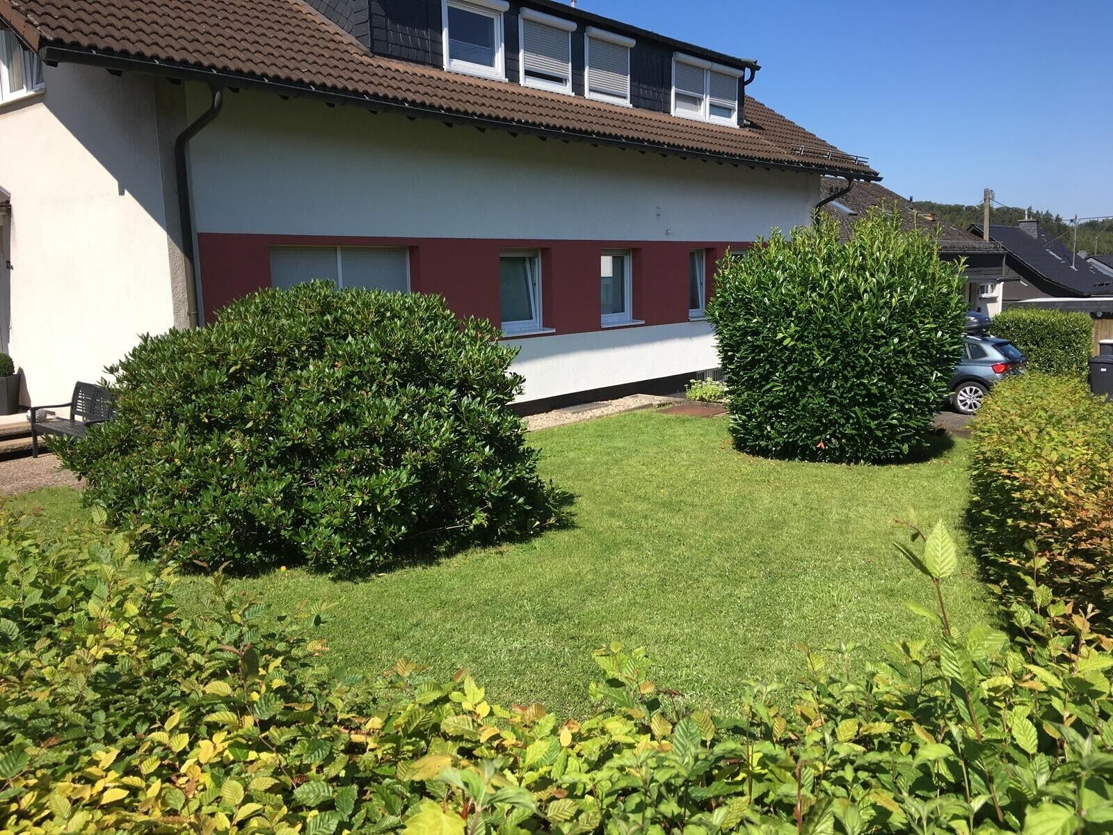 Ein Wohngebäude mit roter und weißer Fassade, zwei gepflegten Hecken und einem ordentlich geschnittenen Rasen.