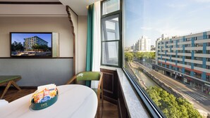 Camera d'albergo con un'ampia finestra che si affaccia su un fiume e degli edifici. Si nota un tavolo da pranzo con una sedia verde e un tavolino con un cesto di oggetti.