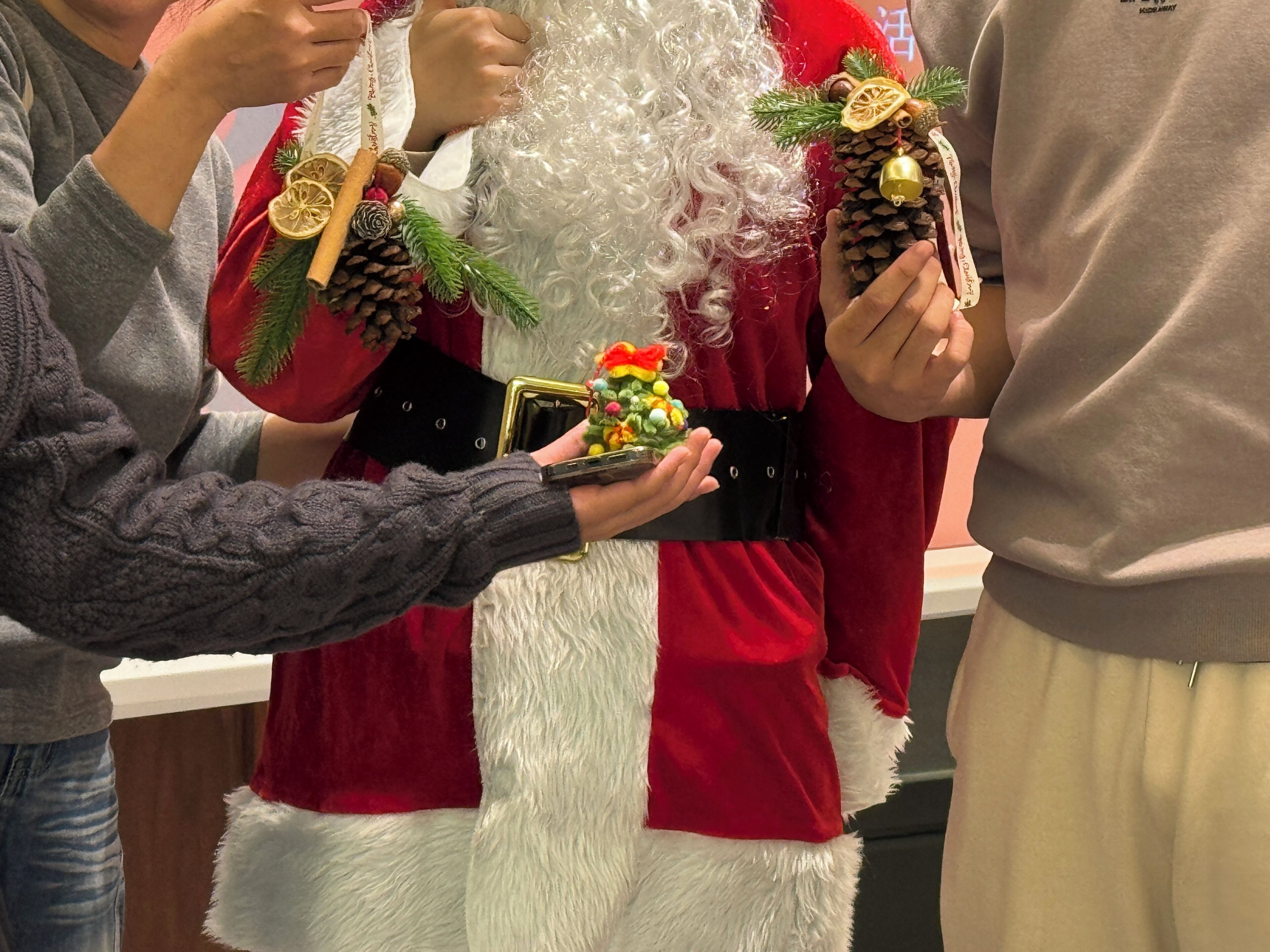 Une personne déguisée en Père Noël, tenant une petite boîte cadeau et une pomme de pin ornée d'une clochette.
