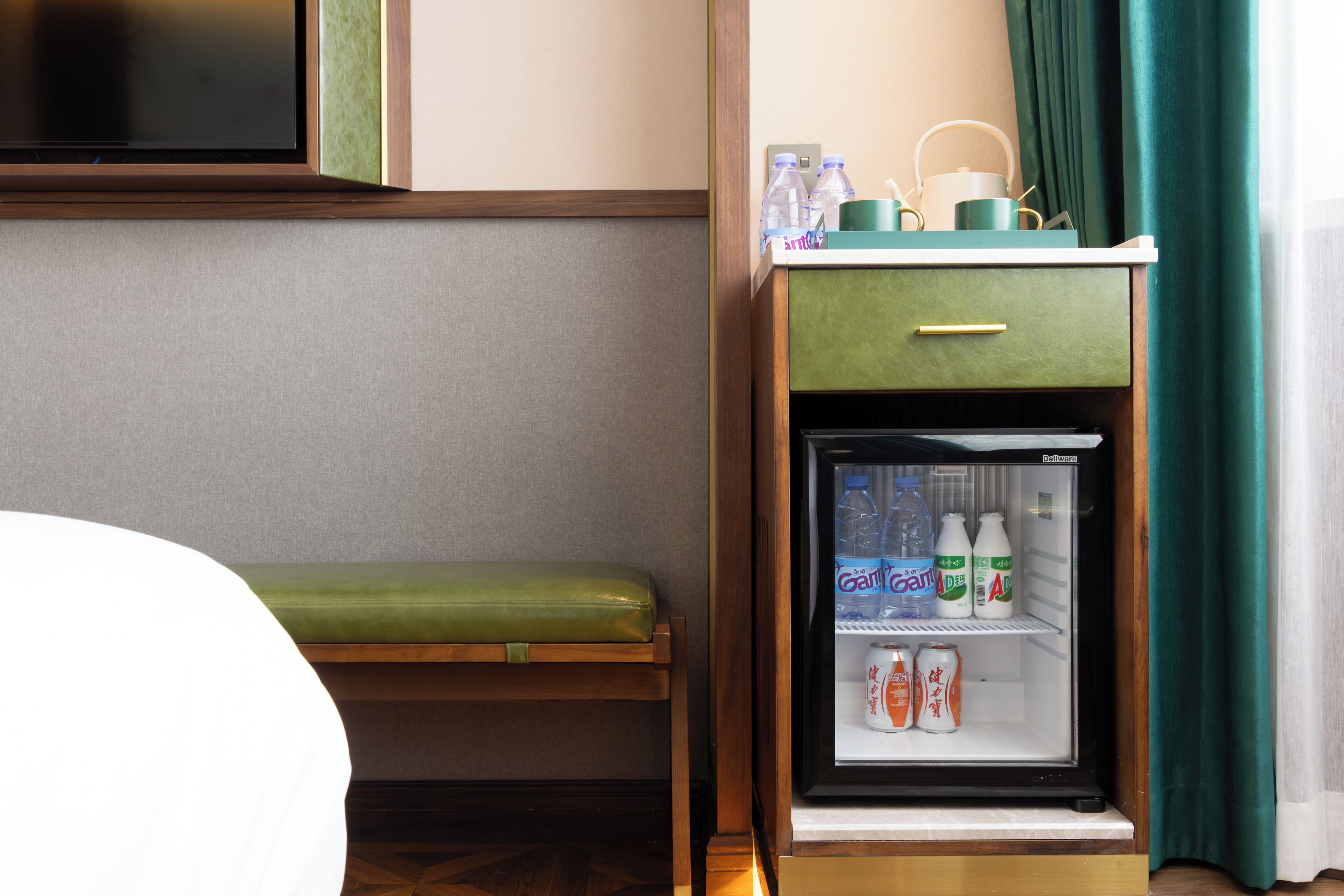 A hotel room with a bed, a green bench, a minibar with beverages, and a small refrigerator.