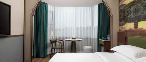 A hotel room with a large bed, a small round table, a chair, a trash bin, and a TV mounted on the wall.