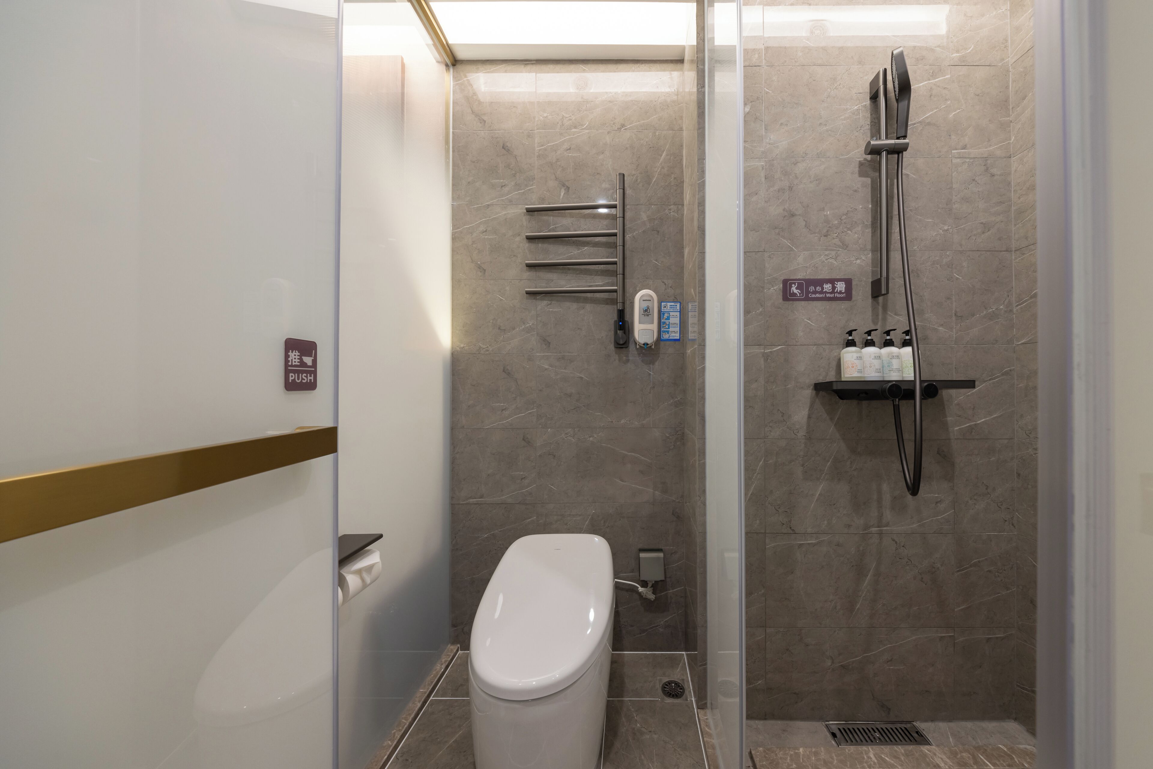 Un baño moderno con inodoro, botón pulsador, secador de manos y zona de ducha con cabezal de ducha manual y grifo empotrado.