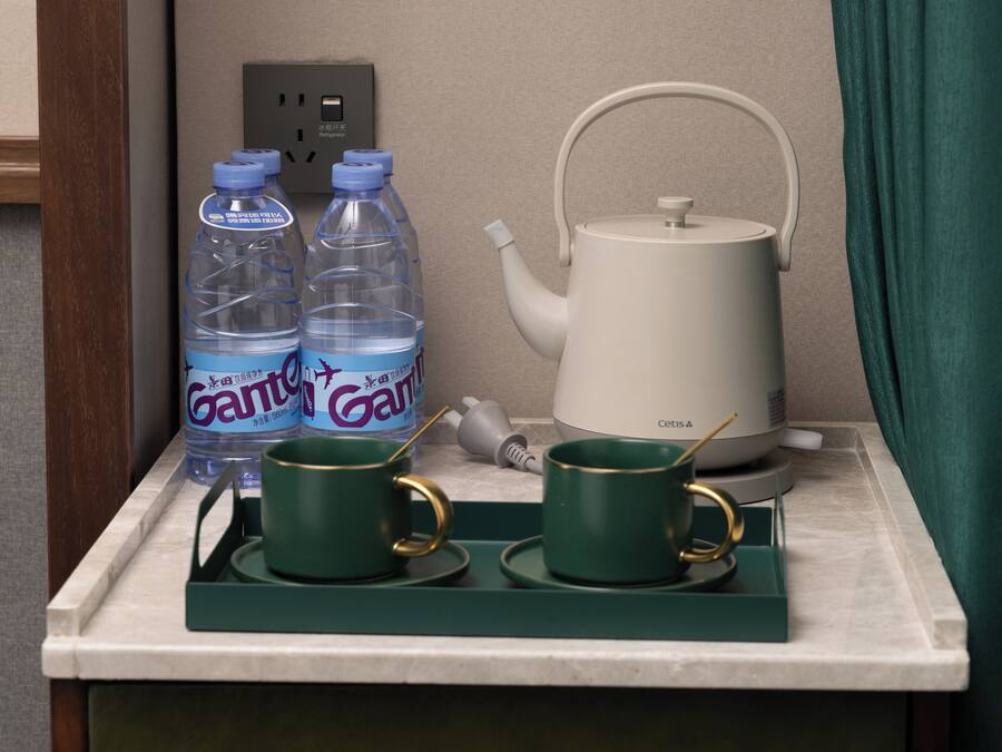 Una encimera de cocina con un hervidor blanco, dos tazas verdes con detalles dorados y dos botellas de agua con tapones azules.