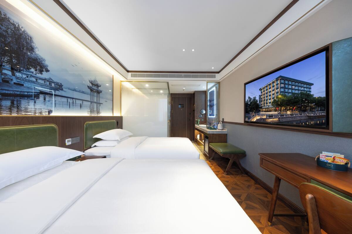A hotel room with two beds, a desk, and a large window with a cityscape view.