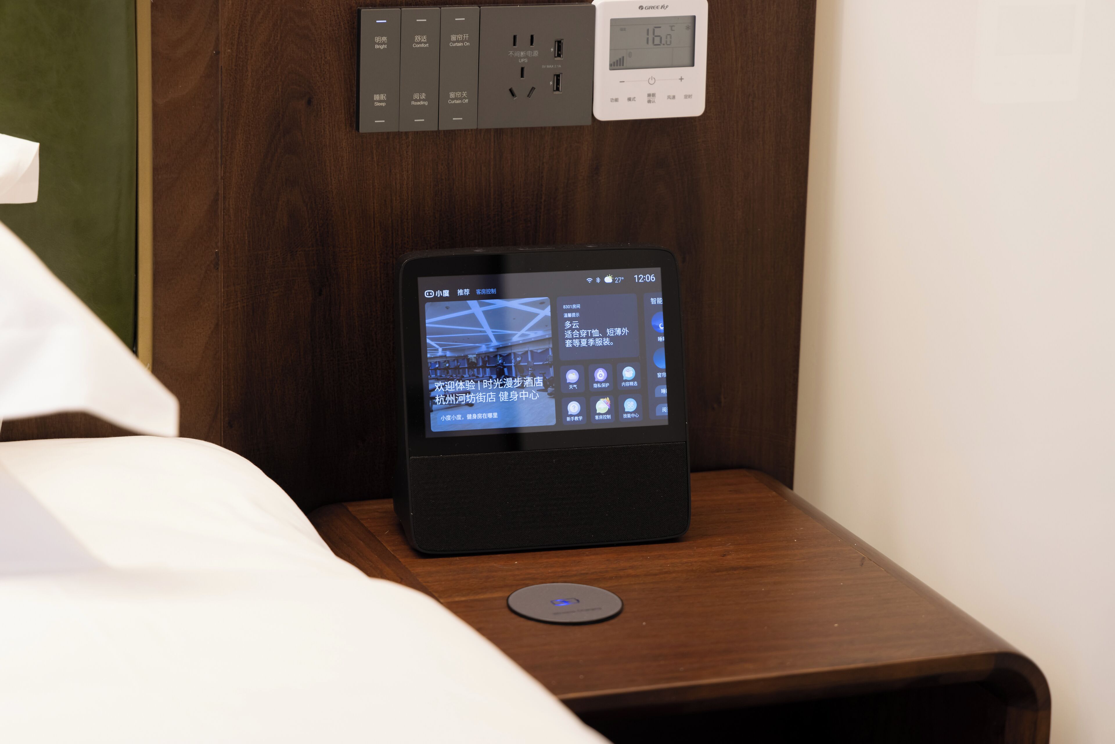 A tablet on a wooden bedside table displaying a digital interface, a power outlet, and a wall-mounted control panel.