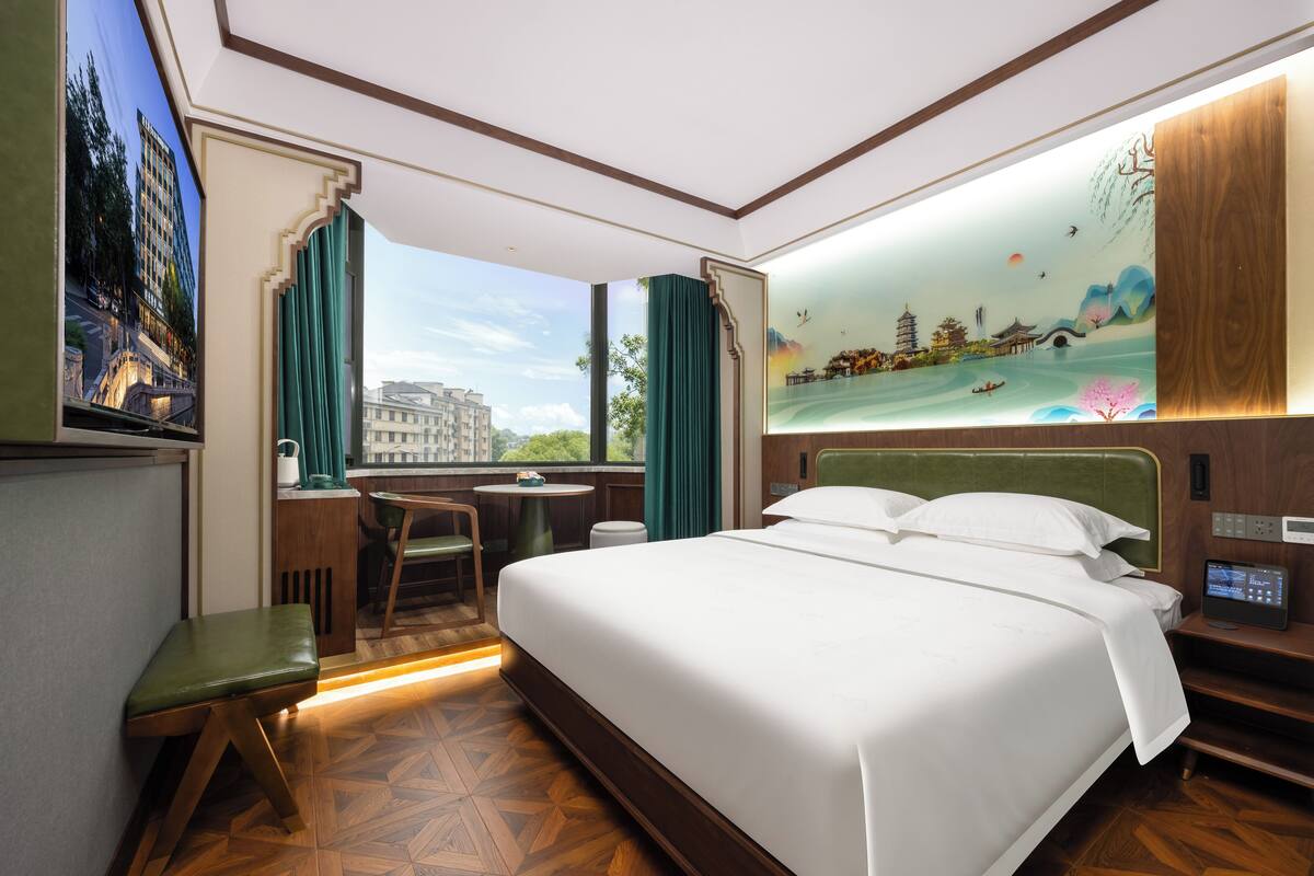 A hotel room with a large bed, a desk, and a view of a cityscape through the window.