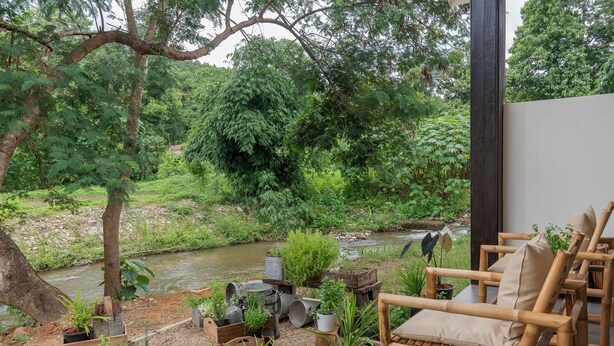 A serene outdoor seating area with wooden furniture and potted plants, overlooking a river and lush greenery.