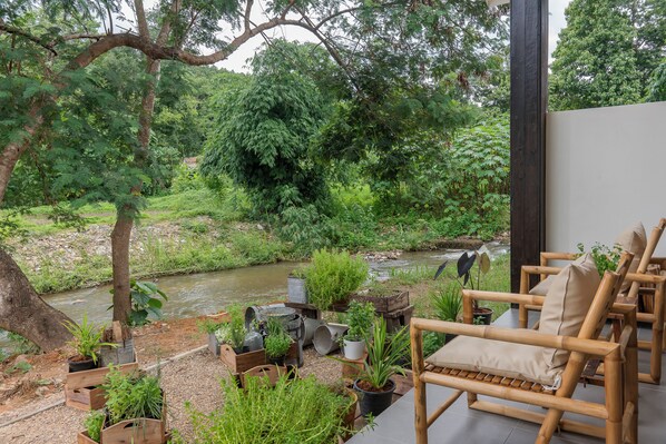 A serene outdoor seating area with wooden furniture and potted plants, overlooking a river and lush greenery.
