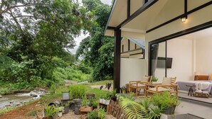 A modern outdoor living area with a view of a stream, surrounded by various potted plants and wooden furniture.