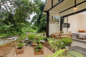 A modern outdoor living area with a view of a stream, surrounded by various potted plants and wooden furniture.
