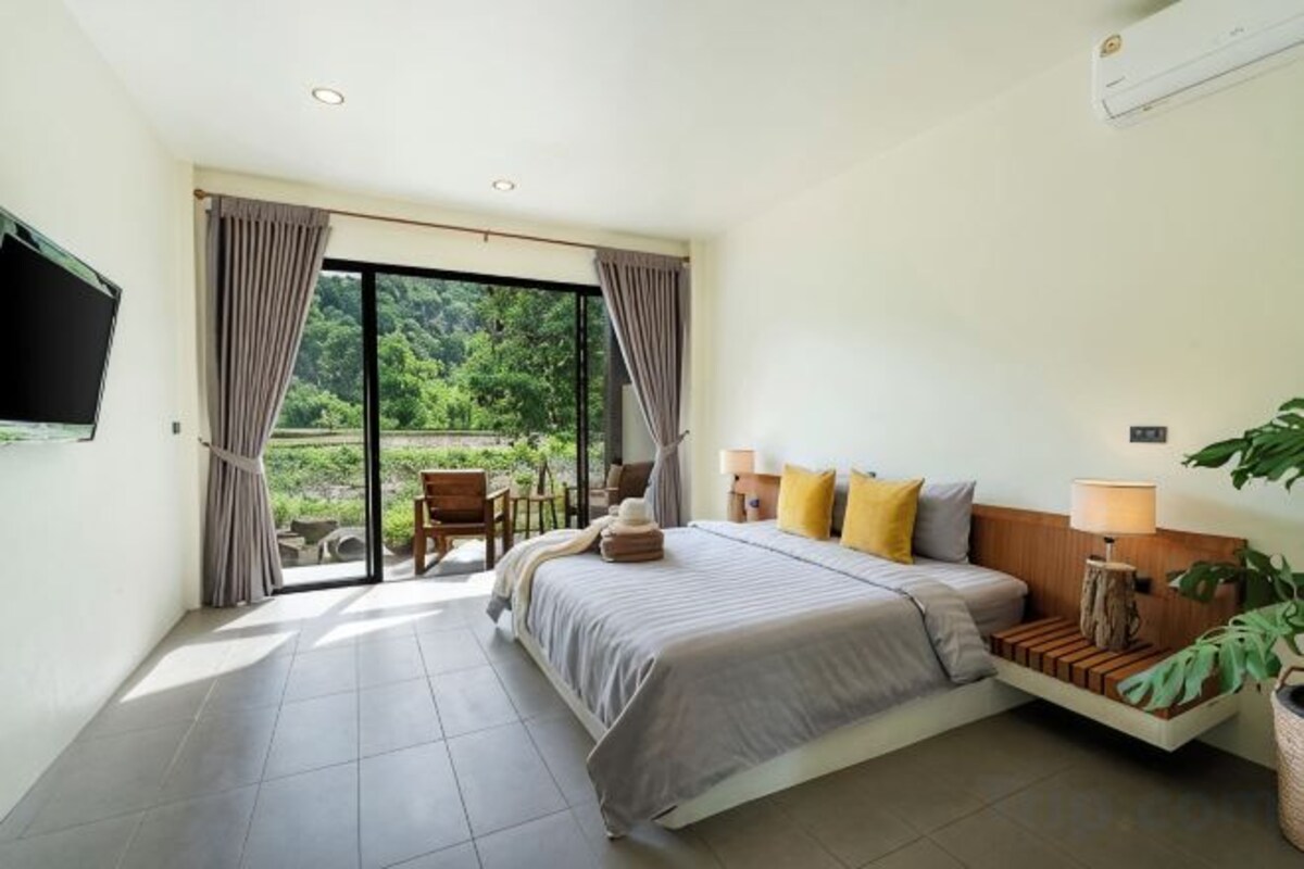 A bedroom with a bed, bedside table, lamp, and a view of greenery through the window.