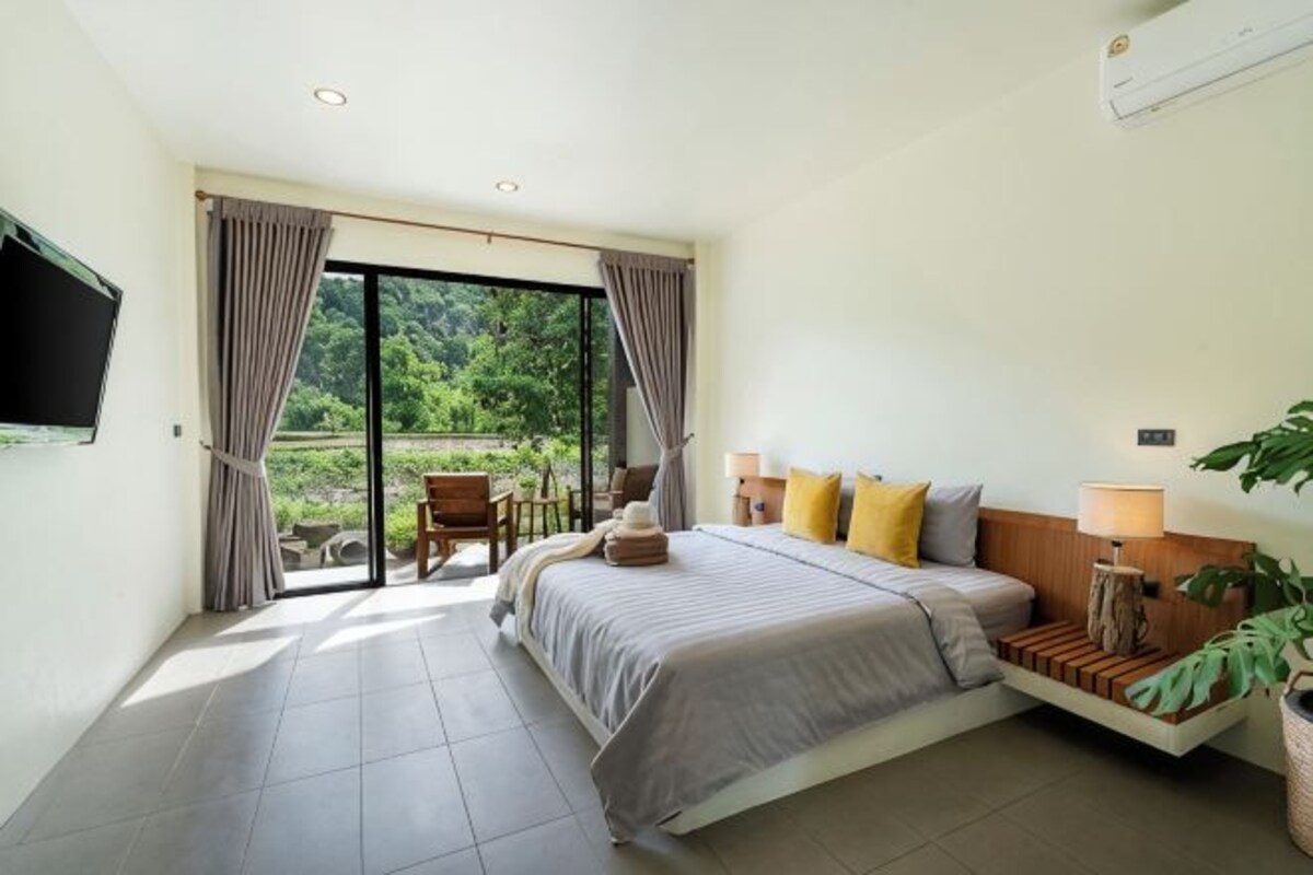 A bedroom with a bed, a bedside table, a lamp, a TV, and a sliding glass door leading to an outdoor area.