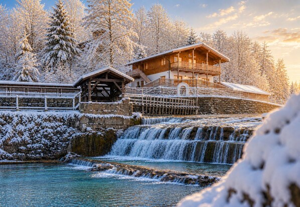 A wooden lodge with a stone water feature and a wooden deck, surrounded by snow-covered trees.