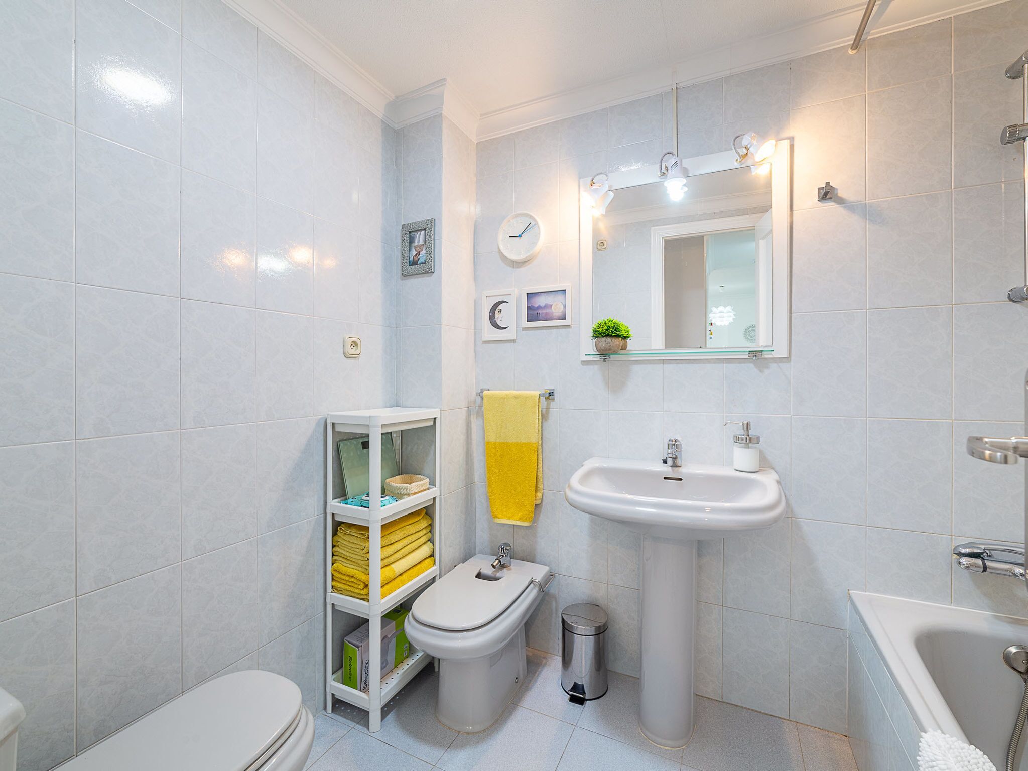 Baño con inodoro, bidé, lavabo y bañera. Se ve una repisa con toallas y un cubo de basura.