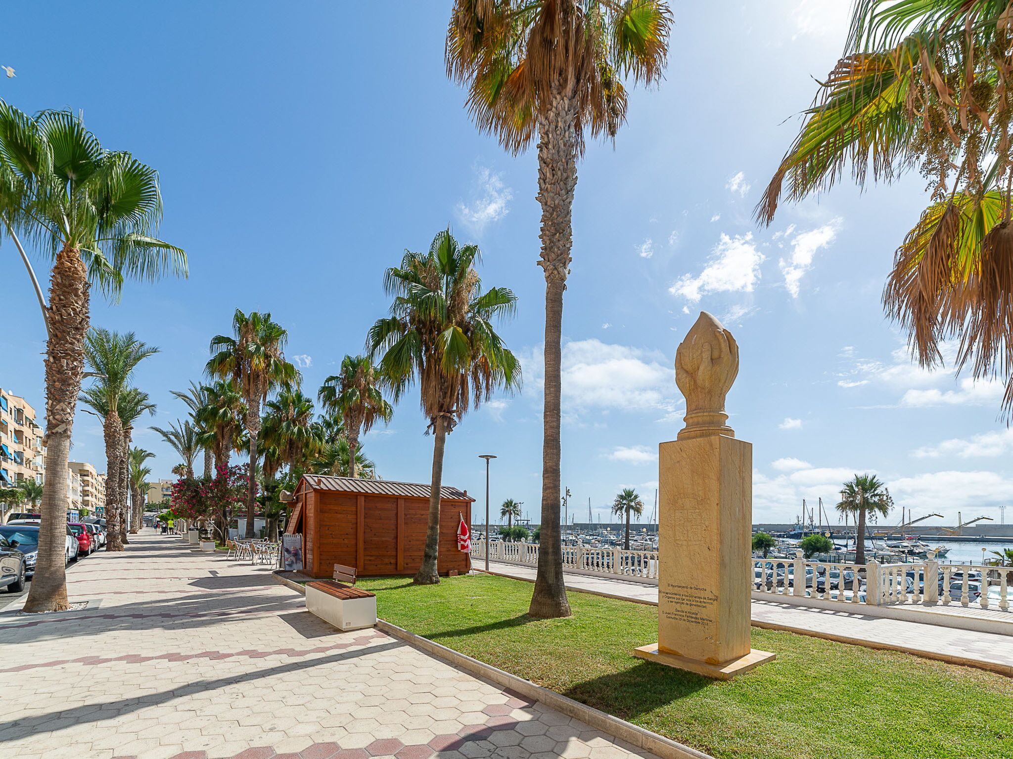 A coastal promenade with palm trees, a wooden structure, and a stone monument.