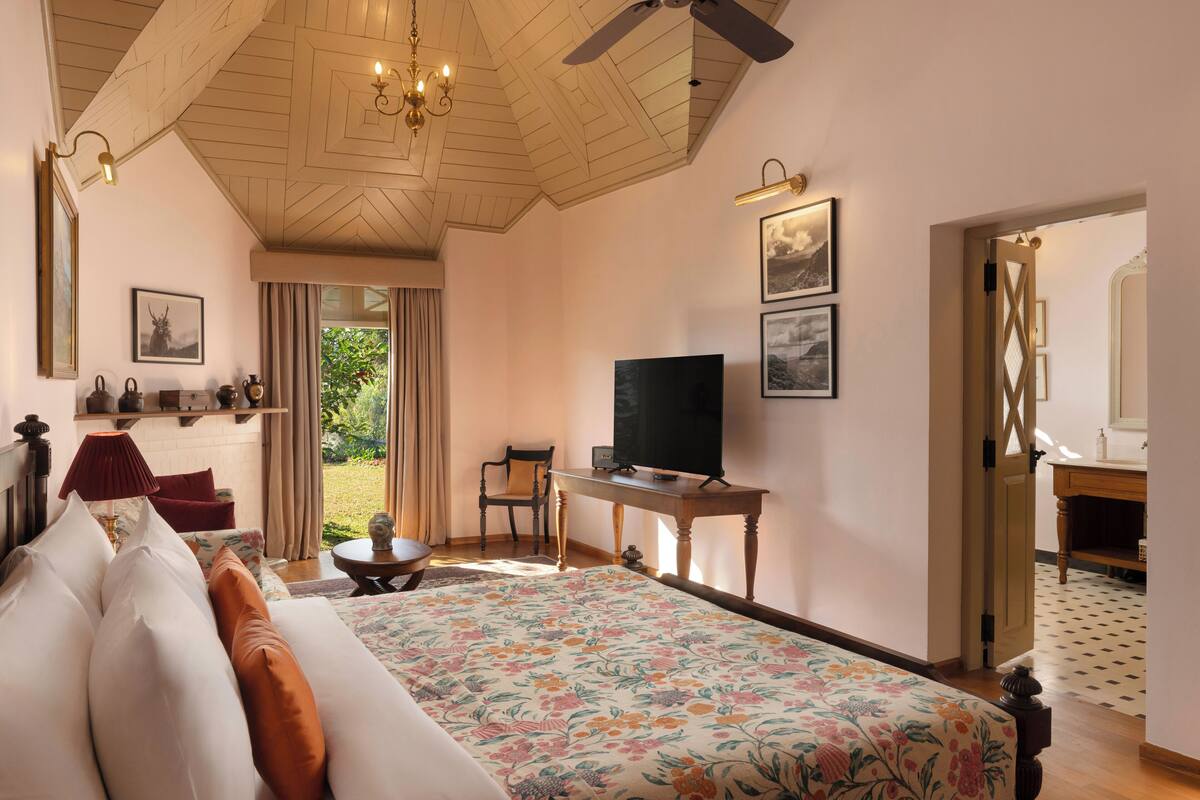 A bedroom with a large bed, a television, a desk, and a chair. There is a window with curtains and a view of greenery outside.