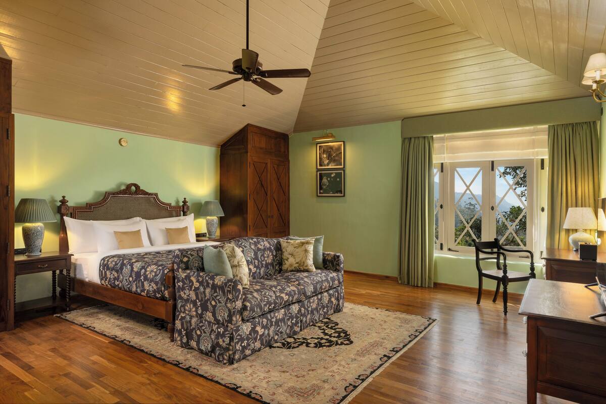 A bedroom with a bed, a sofa, a desk, and a chair. There is a ceiling fan, a window with curtains, and a lamp on the desk.