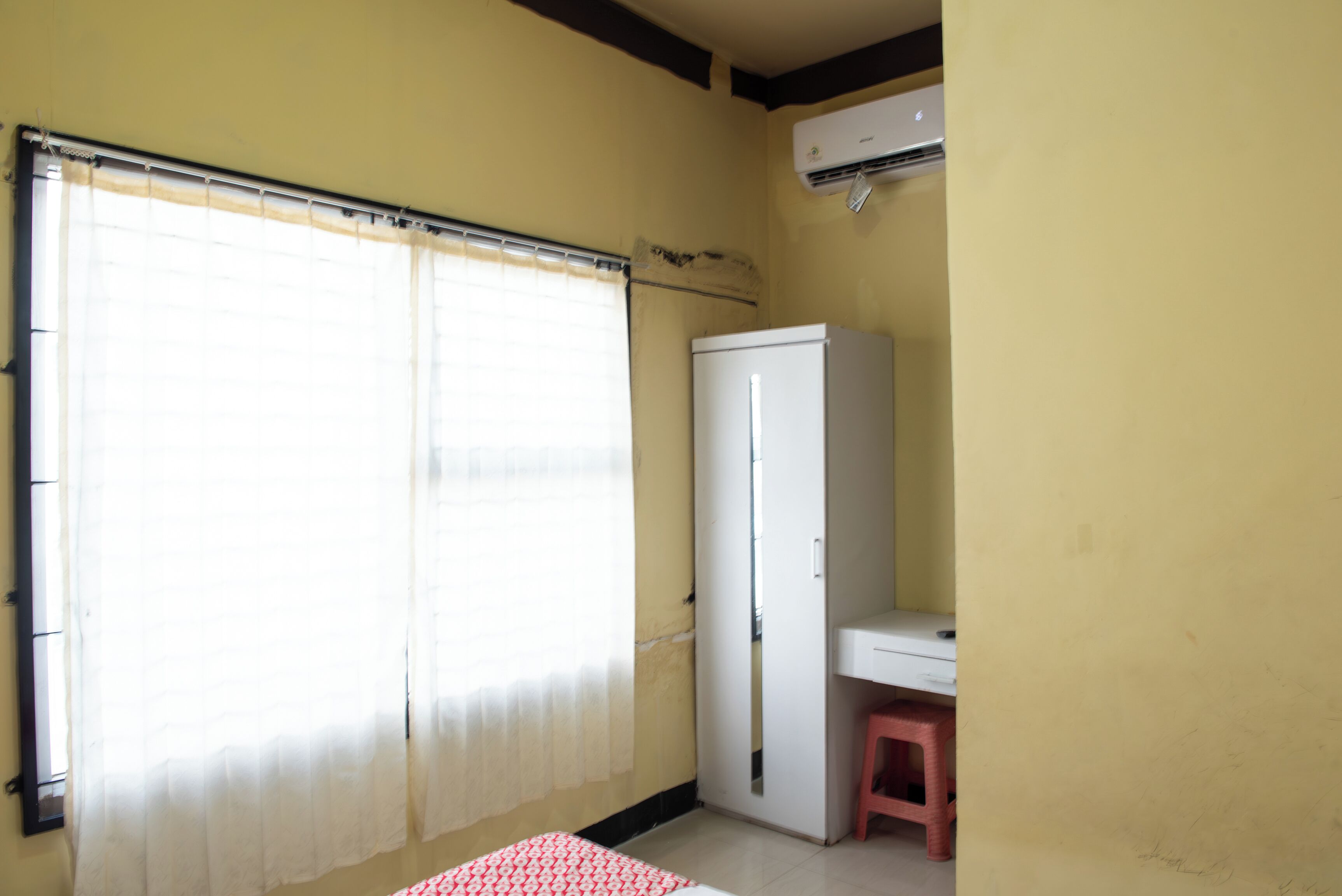 Ein Zimmer mit einem großen Fenster, das mit durchsichtigen Vorhängen bedeckt ist, einem weißen Kleiderschrank, einem kleinen Schreibtisch mit einem pinken Hocker und einer an der Wand montierten Klimaanlage.