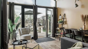 A modern living room with a sofa, a coffee table, and a dining table. There is a large sliding glass door leading to an outdoor patio area with a grill and umbrellas.