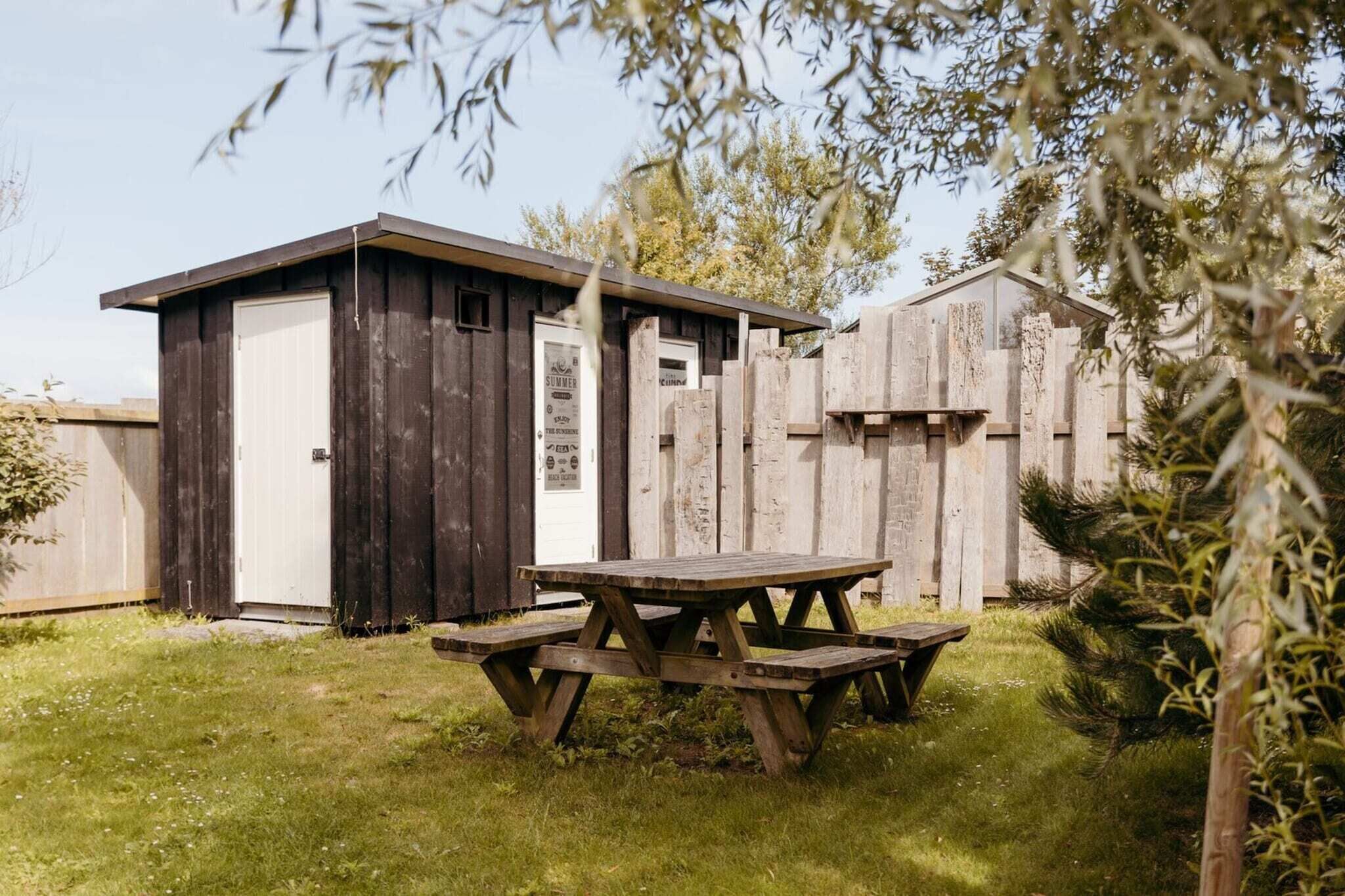 Un abri en bois avec une porte et une enseigne, une table de pique-nique et une clôture en bois, le tout situé dans une cour verdoyante.