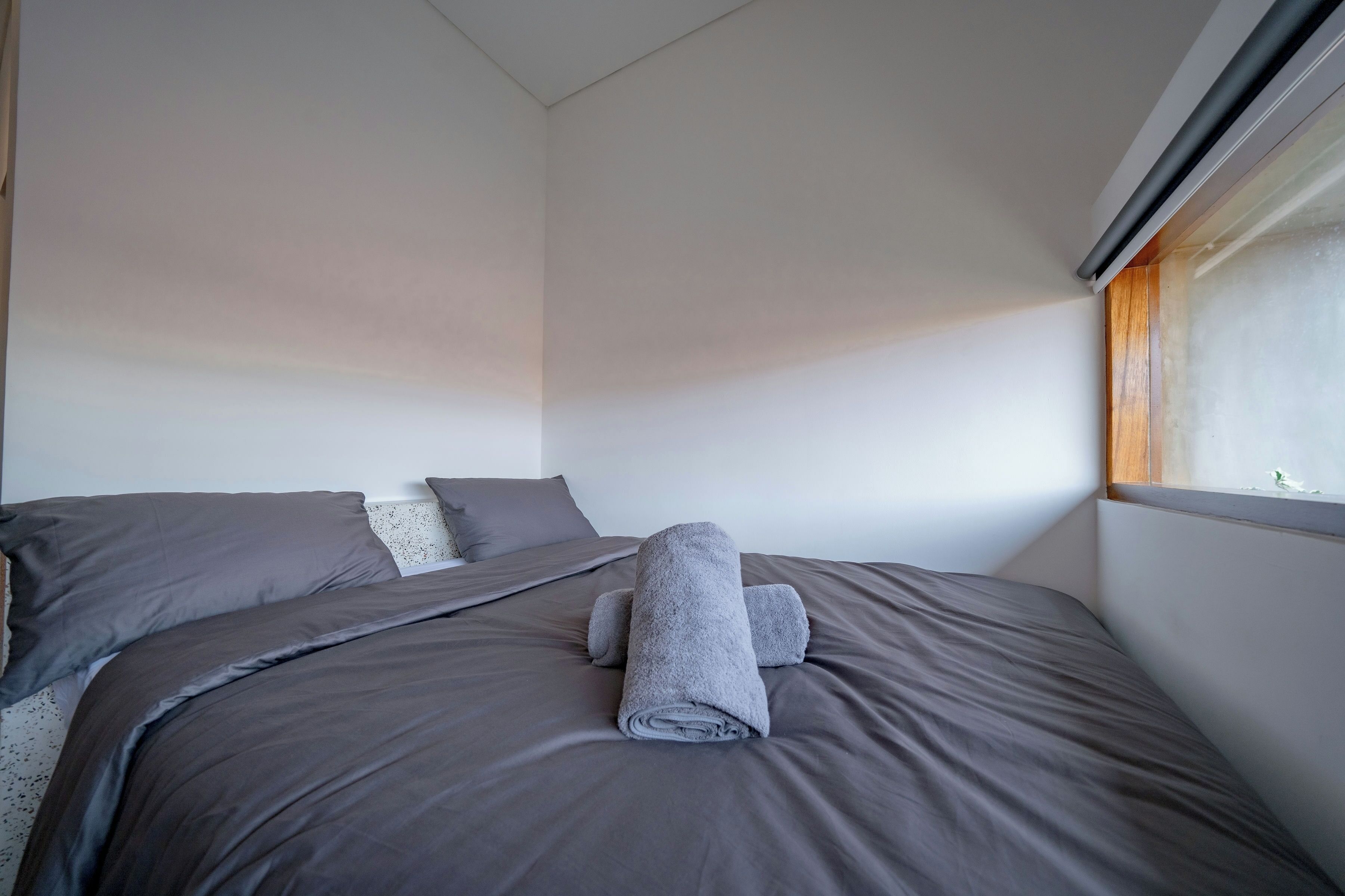 Una cama bien hecha con ropa de cama gris y toallas enrolladas, una ventana con persianas, y una pared lisa.