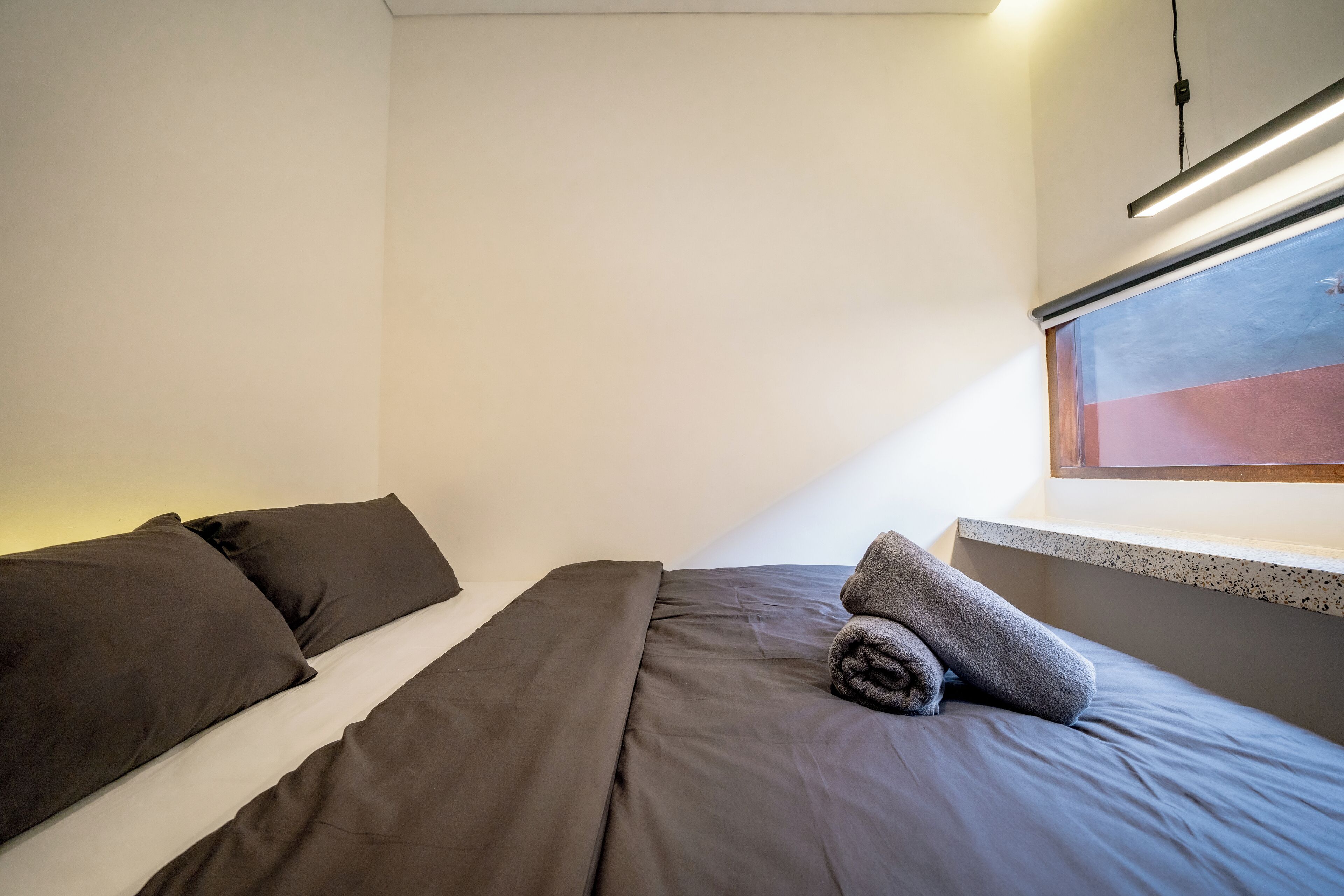 Un lit simple avec une literie sombre et une serviette enroulée, une petite fenêtre donnant sur le paysage et un luminaire au plafond.