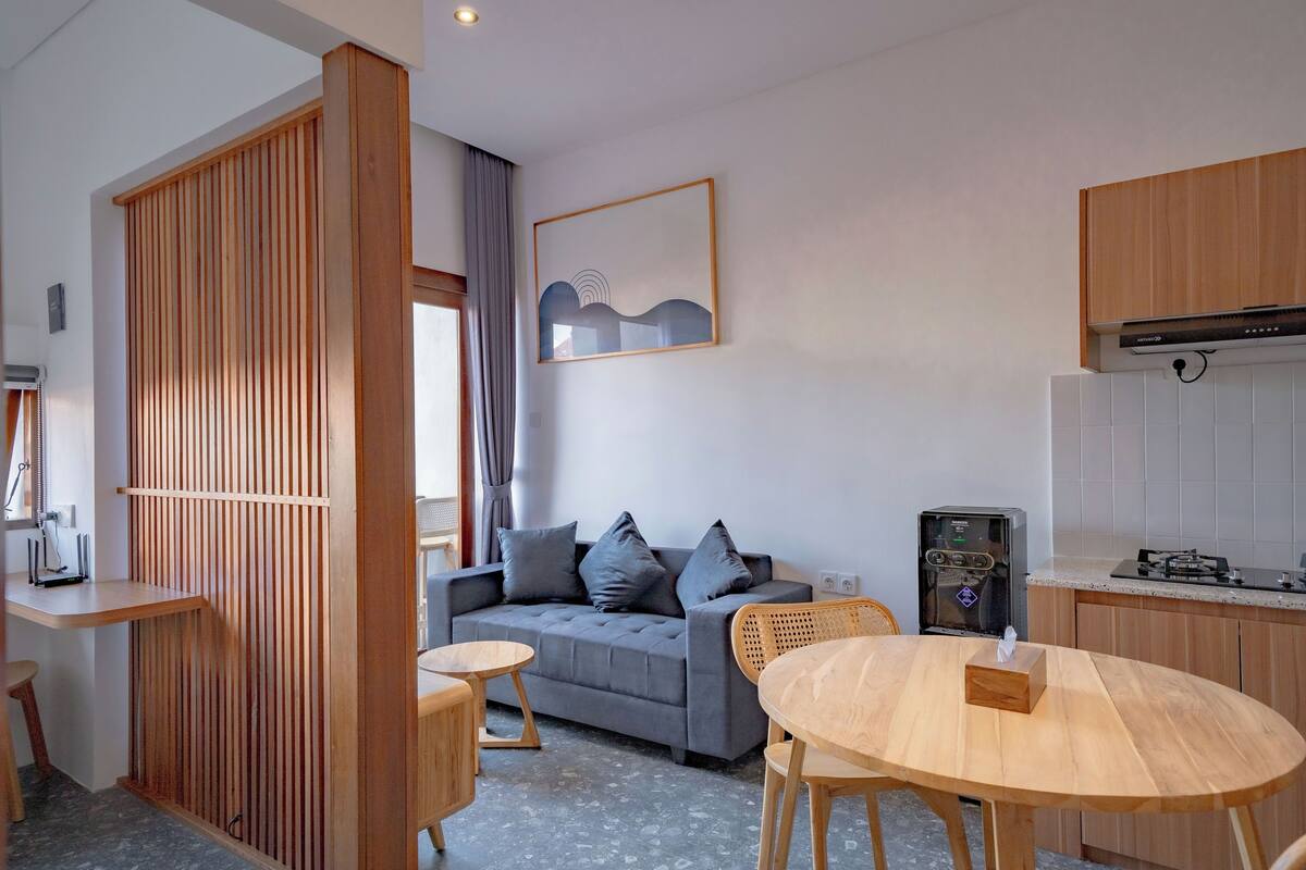 Ein modernes Wohnzimmer mit grauem Sofa, Holzelementen, einem Esstisch und einer Küchenzeile mit Wasserspender.