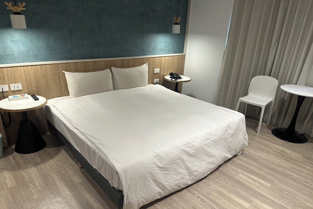 A hotel room with a large bed, a bedside table with a phone, a chair, a small table, and a curtain.