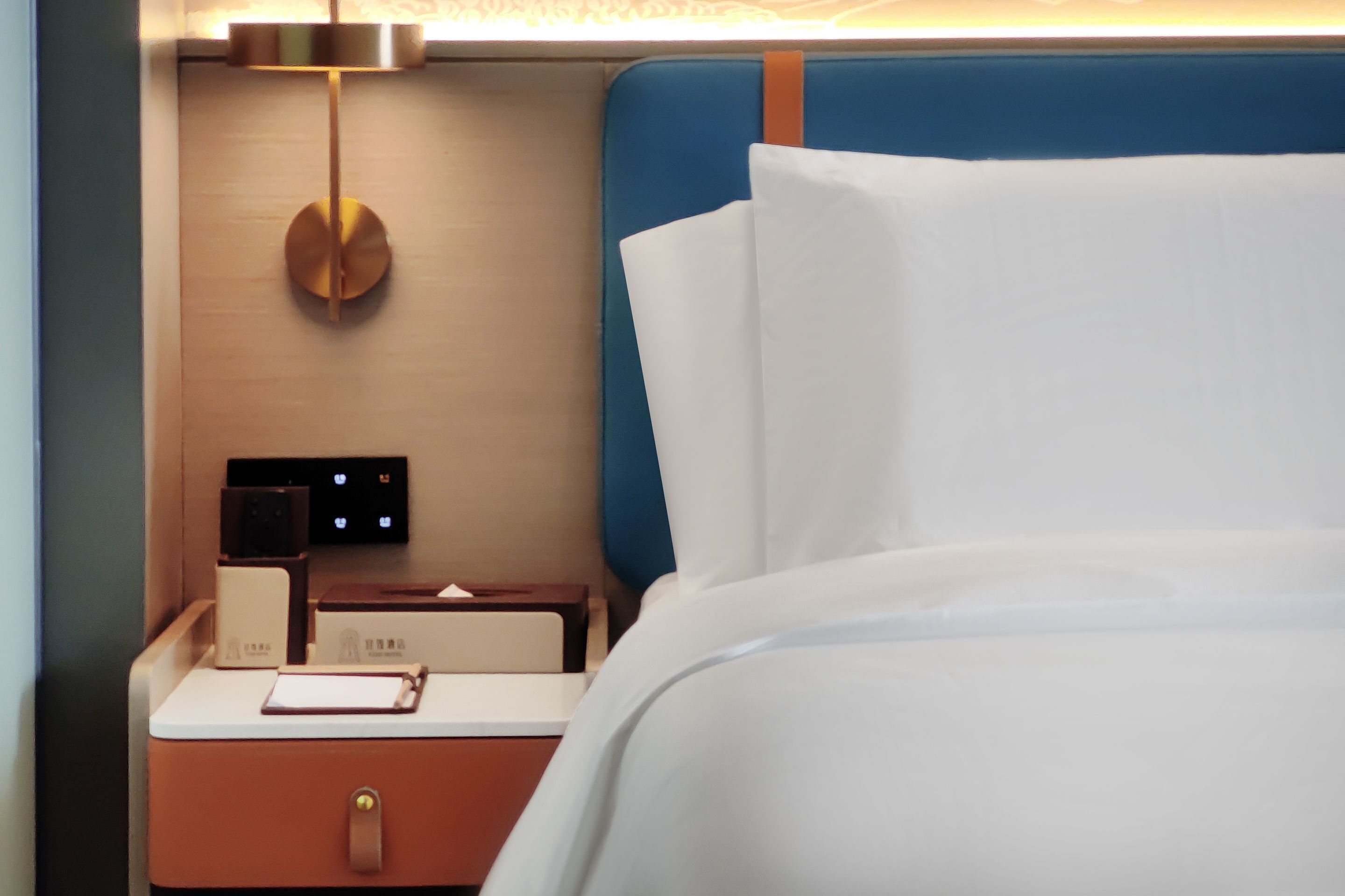 Una cama bien hecha con sábanas blancas, una mesita de noche con una lámpara, un reloj y un pequeño estuche.