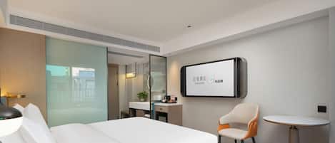 Una habitación de hotel moderna con una cama grande, una pequeña mesa redonda, una silla, un televisor montado en la pared y un baño visible a través de una puerta de cristal.