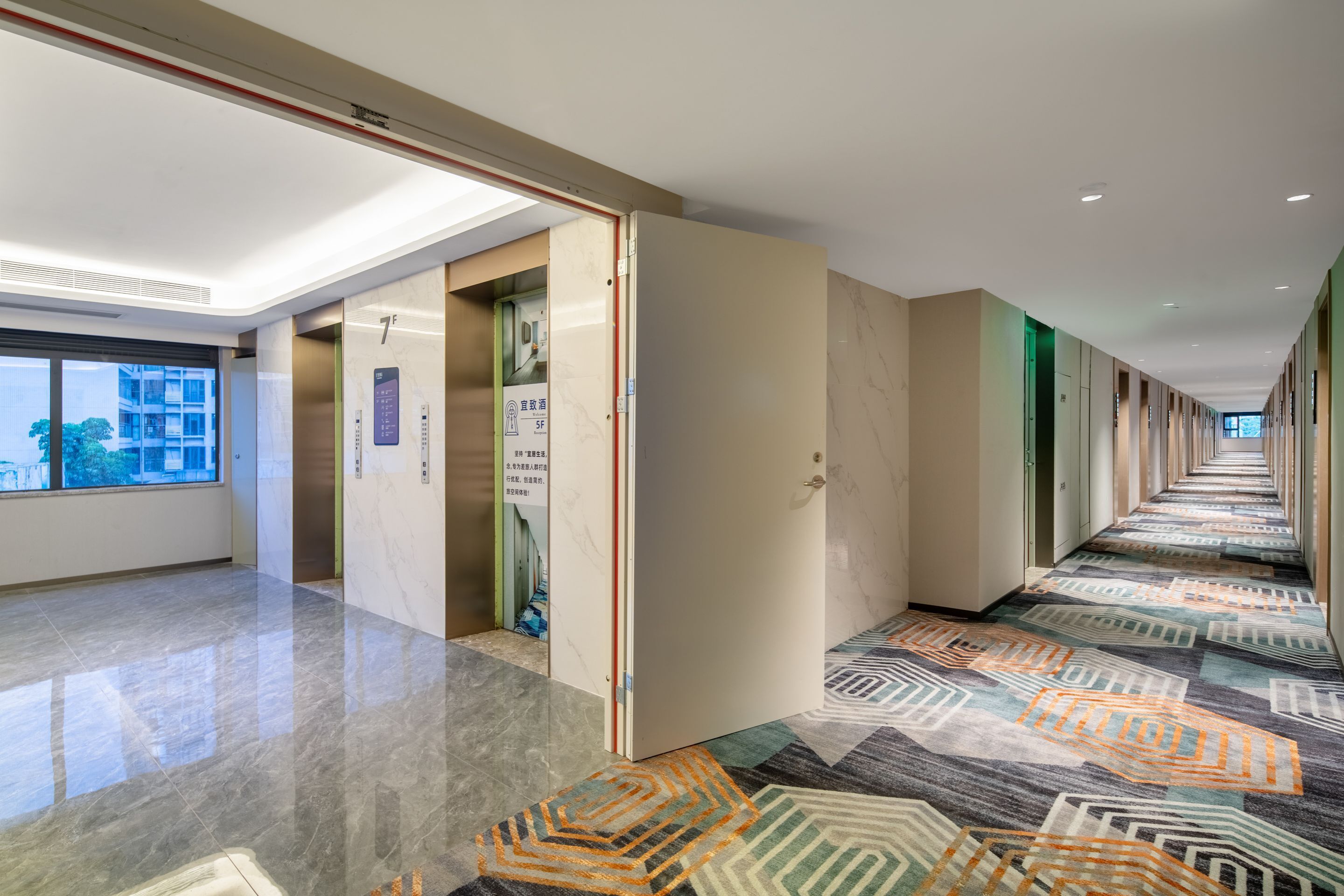 Un pasillo de hotel con alfombra estampada, una puerta de vidrio que da a un ascensor y vistas al exterior por una ventana.