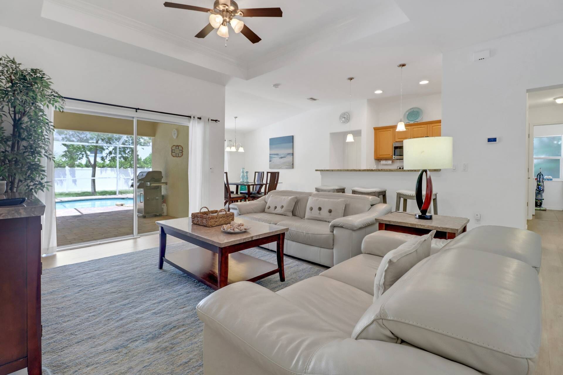 A spacious living room with a beige sofa, a coffee table, and a ceiling fan. There is a dining area with chairs and a table, and a kitchen area with a lamp and a countertop.