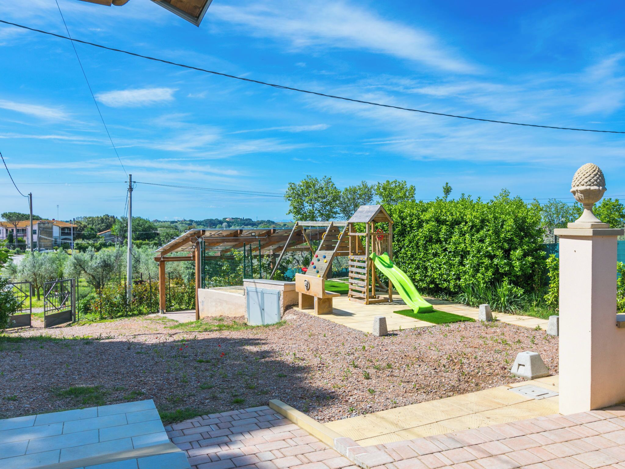 A playground with a slide and climbing structures, surrounded by greenery and a clear sky.