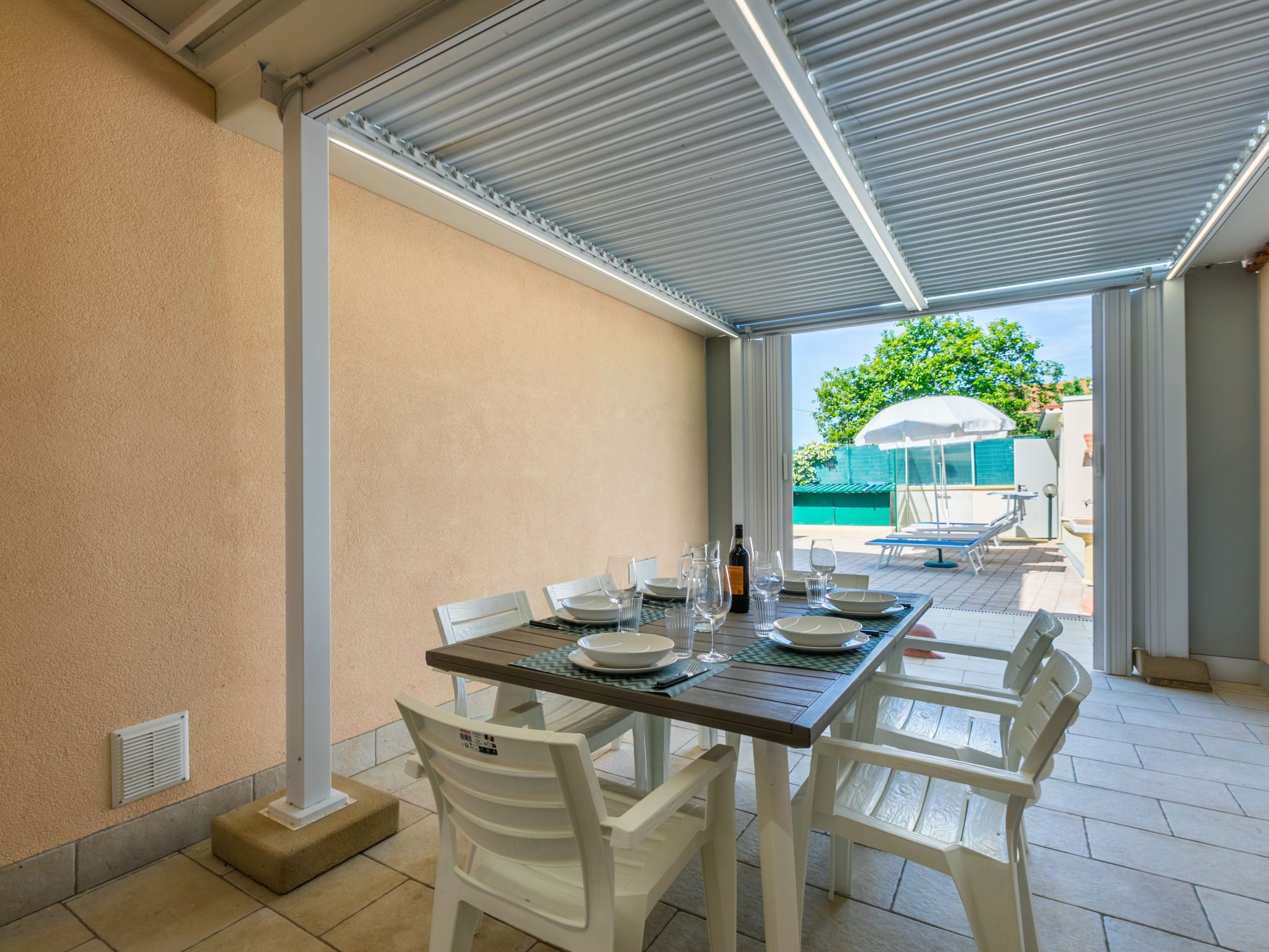 A covered patio with a table set for dining, white plastic chairs, and a view of a swimming pool and greenery in the background.
