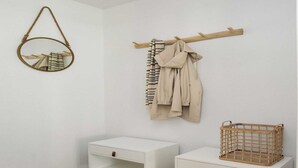 Une pièce minimaliste avec un mur blanc, un porte-manteau en bois sur lequel est accroché un manteau beige, un miroir rond et une table de chevet blanche sur laquelle se trouve un panier en osier.