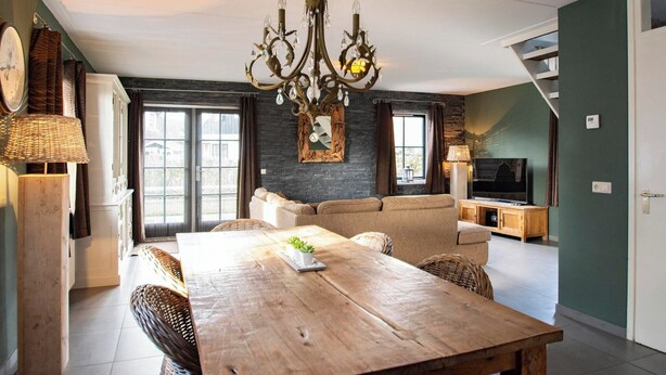 A dining area with a wooden table, wicker chairs, a chandelier, and a television in the background. - Villa in Callantsoog with Whirlpool and Beach (Callantsoog)