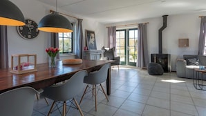 A dining area with a wooden table, grey chairs, and pendant lights. There is a fireplace, a sofa, and a large window with curtains.